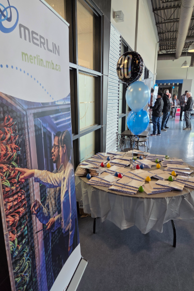 Image of a MERLIN banner placed at the entrance of an event. A table with notebooks and balloons is visible in the background with attendees.