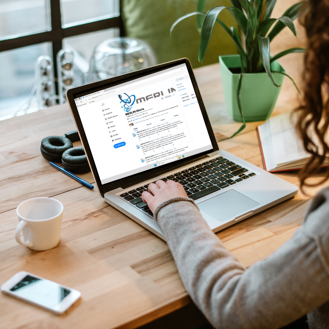 Image of a desk with a laptop, coffee cup, smart phone, and plant. A persons arm and back of head is visible with their arm resting on the laptop keyboard. The screen is showing the MERLIN BlueSky Alerts account