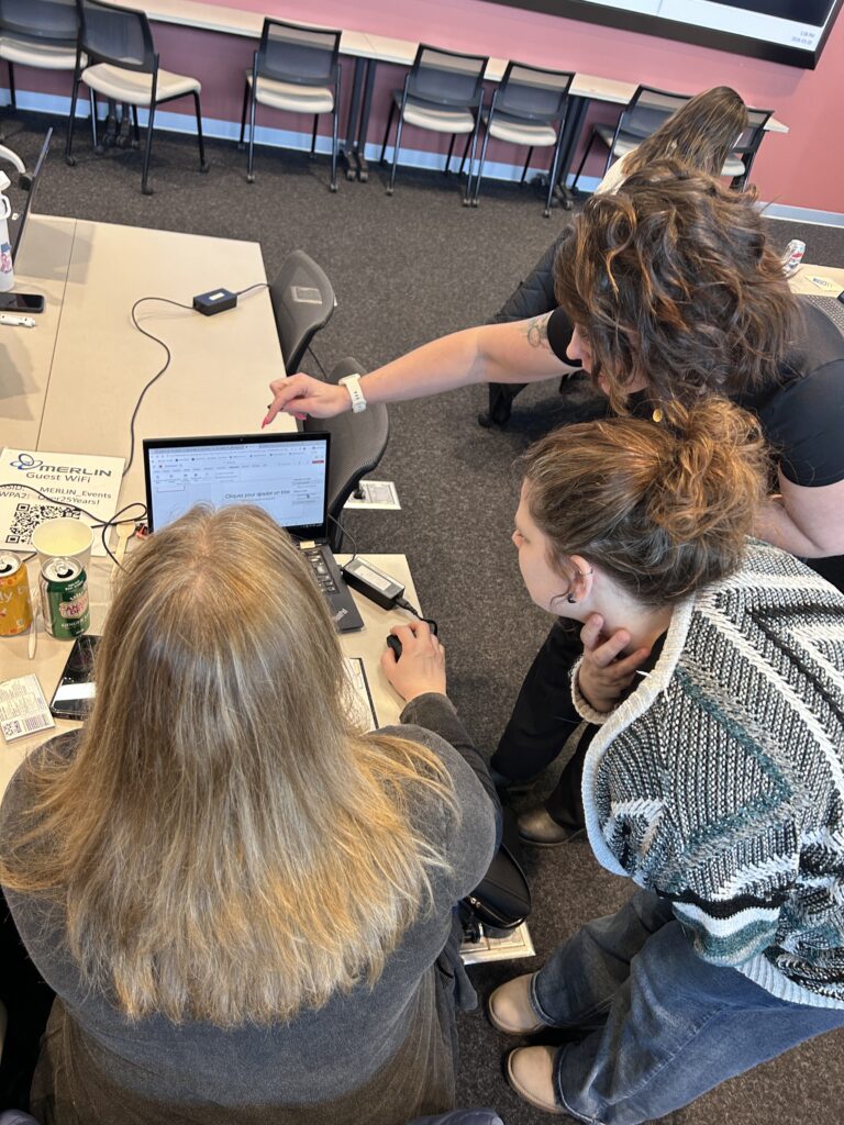 3 individuals, viewed from the back, looking at a computer displaying Microsoft accessibility tools, one person is pointing at the screen