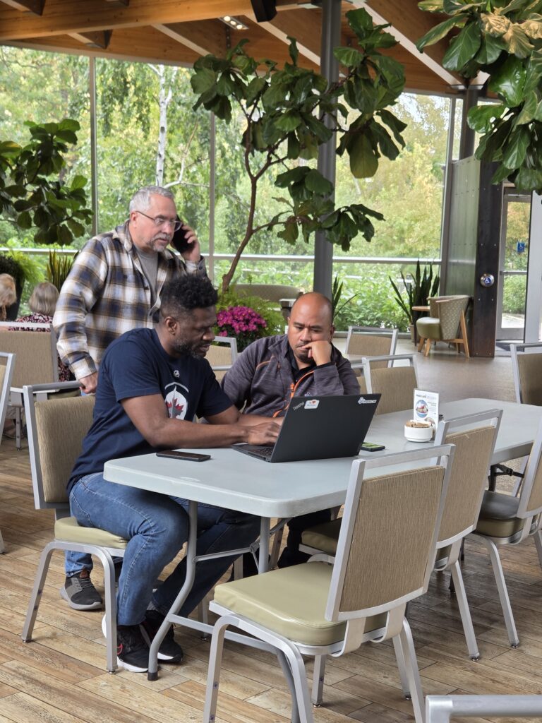 3 people working at a laptop. Two seated and one standing behind on the phone
